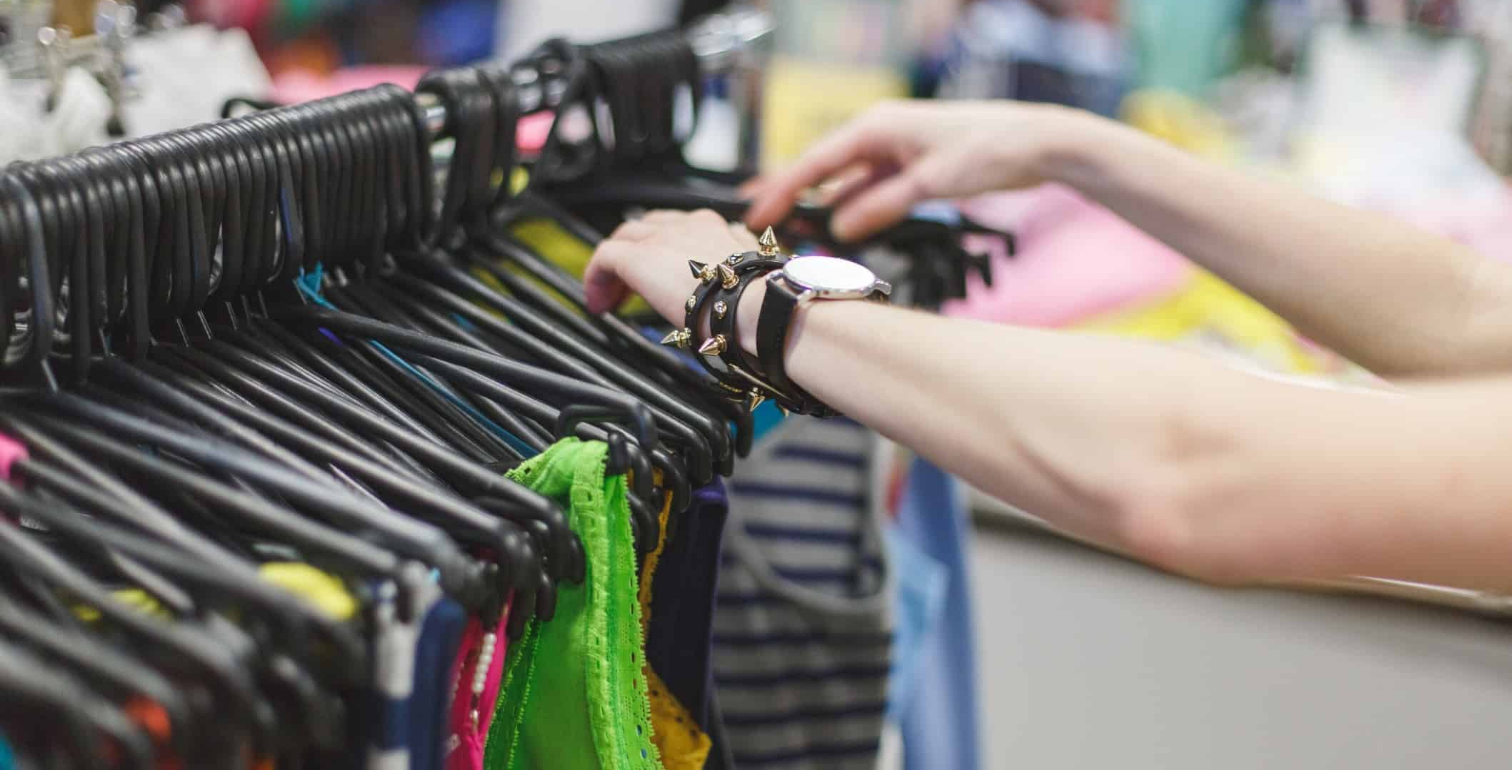 mãos de um jovem percorrem um cabide de chão cheio de cabides individuais de roupa, exposta em ambiente de feira