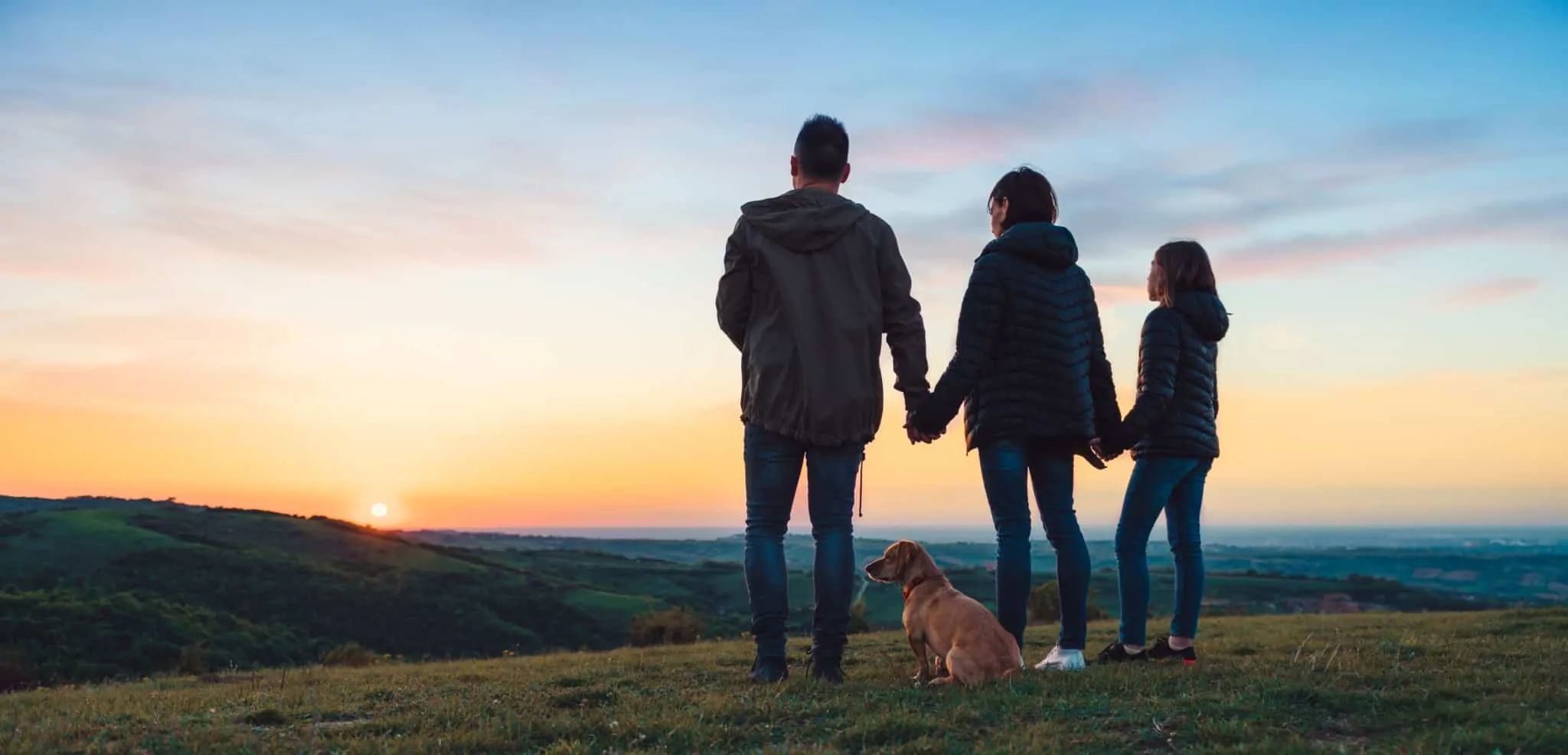 Autorização de residência: como obter, quais os direitos e obrigações Uma família (pai, mãe e criança) de mãos dadas a ver o pôr do sol, acompanhados pelo cão