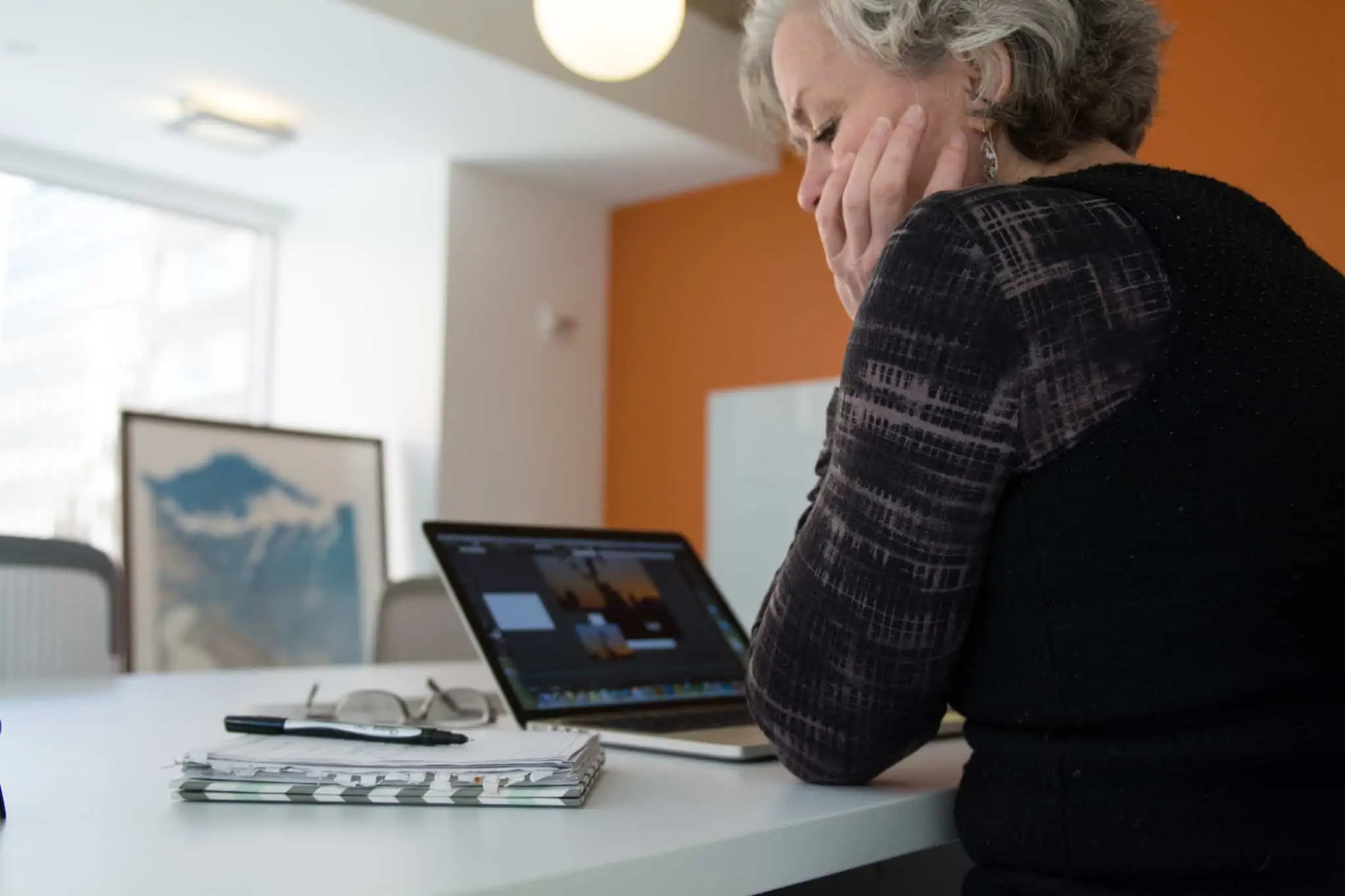 Mulher de cabelo grisalho consulta uns apontamentos num caderno, enquanto trabalha num computador