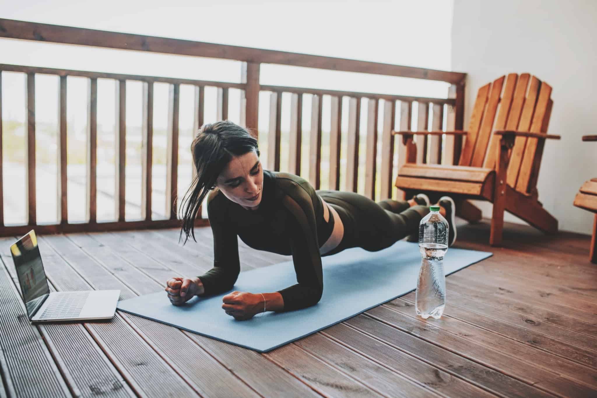 Mulher faz exercício físico na varanda, em frente ao um computador portátil