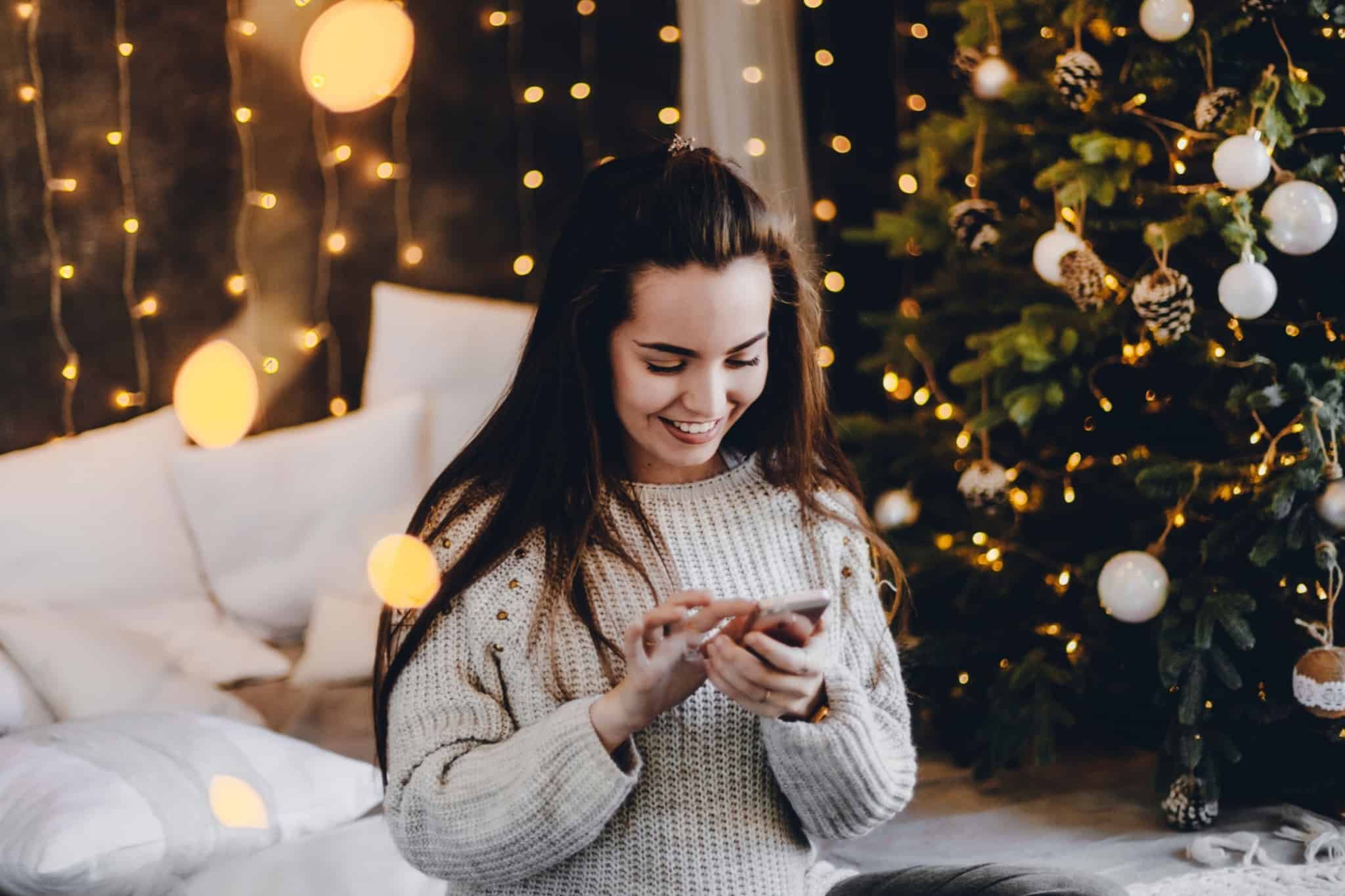 10 dicas para passar uma consoada em época Covid Rapariga a sorrir, com o telemóvel na mão e a árvore de Natal iluminada atrás