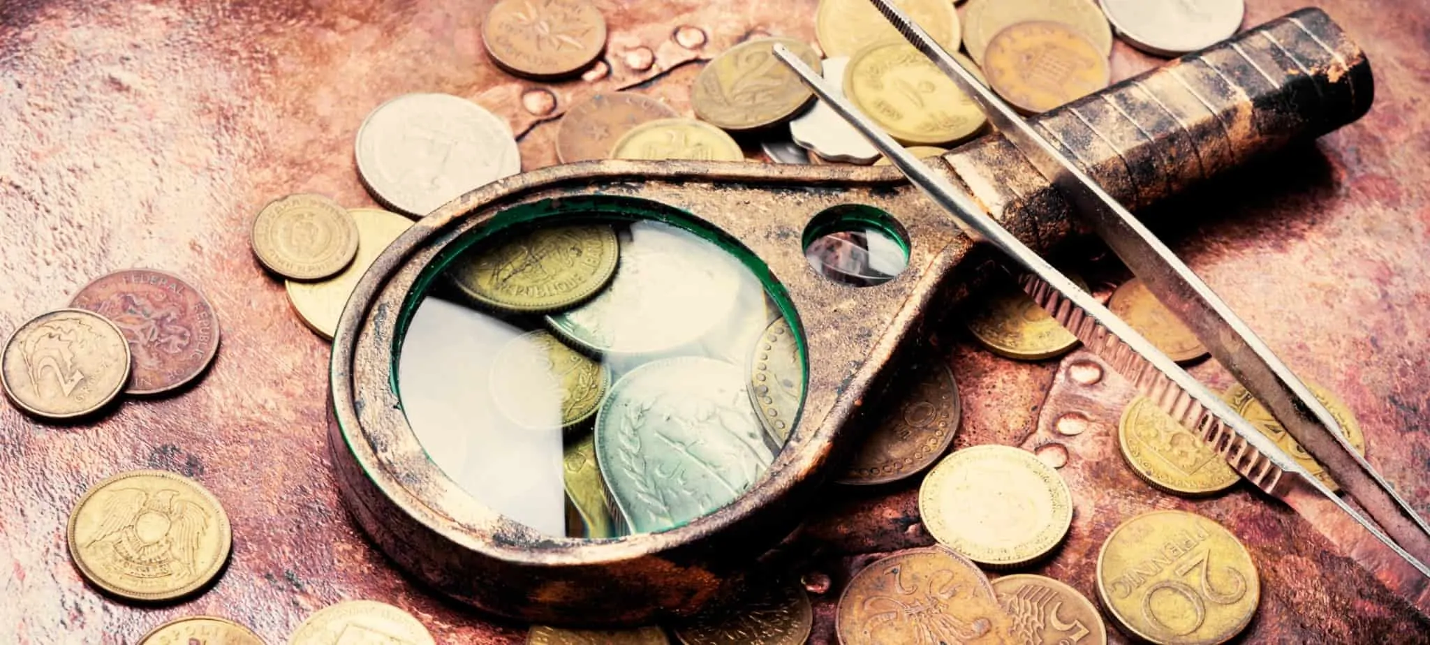 Onde posso vender as minhas moedas antigas? Várias moedas com aspeto antigo sobre uma mesa de madeira. Em cima uma lupa e uma pinça