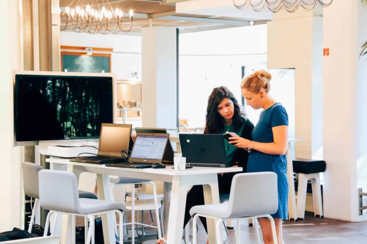 Duas mulheres conversam junto a uma mesa alta em frente a computadores e aos telemóveis