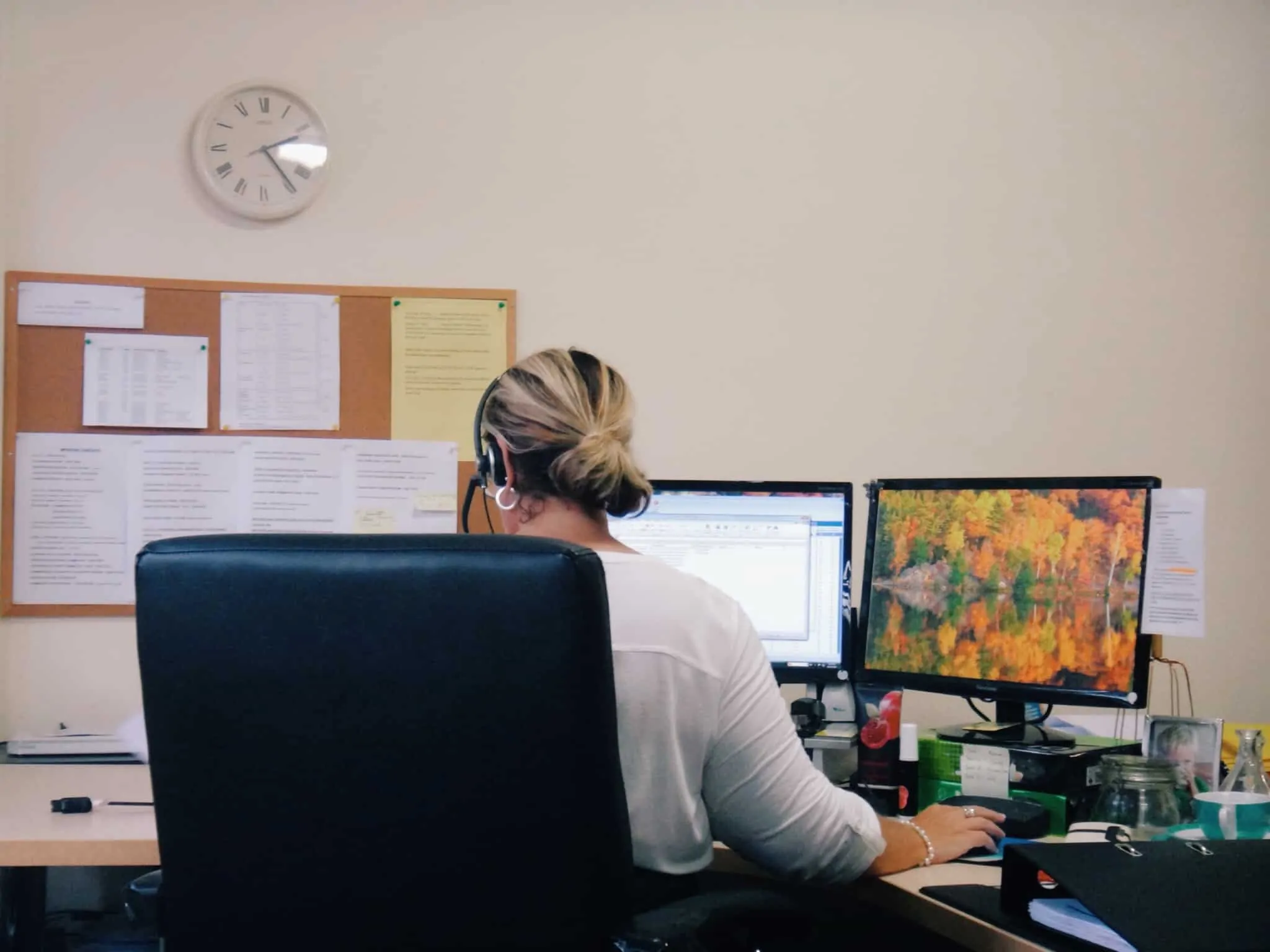 Regime de adaptabilidade laboral: Como funciona, quais os direitos e deveres TRabalhadora sentada à secretária, em frente ao computador. Em frente tem um placar com folhas penduradas e um relógio de parede
