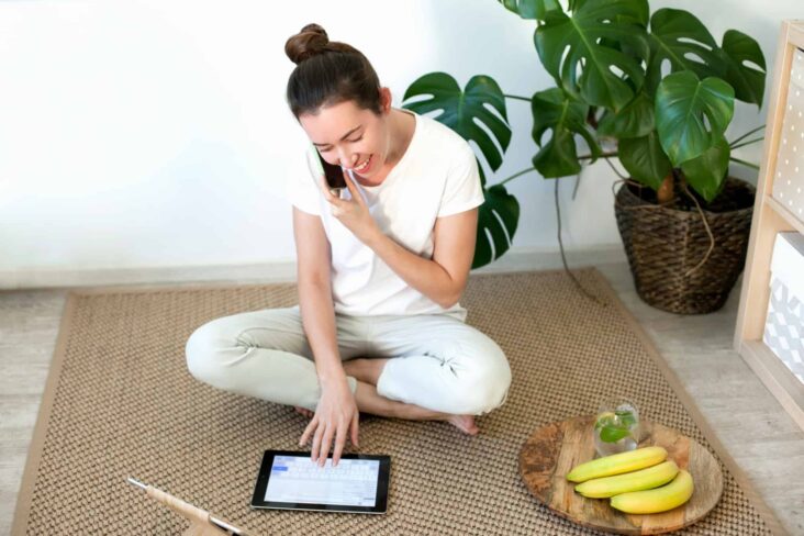 Mulher sentada no chão de casa a falar ao telemóvel ao mesmo tempo que consulta um tablet. À sua frente tem um prato com bananas