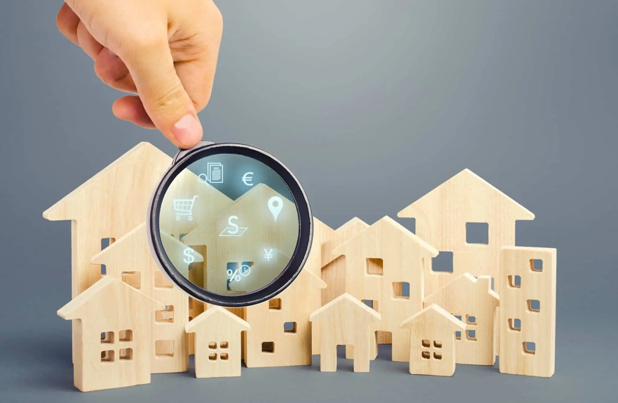Vai vender casa? Saiba quais os custos associados Figuras de casas e prédios em madeira e uma lupa à frente através da qual se veem simbolos de dinheiro, localização e compras