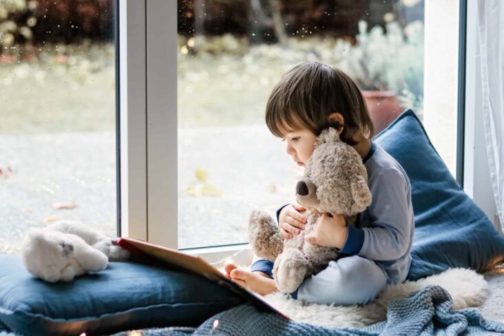 Criança está junto a uma janela a ver um livro enquanto agarra um urso de peluche
