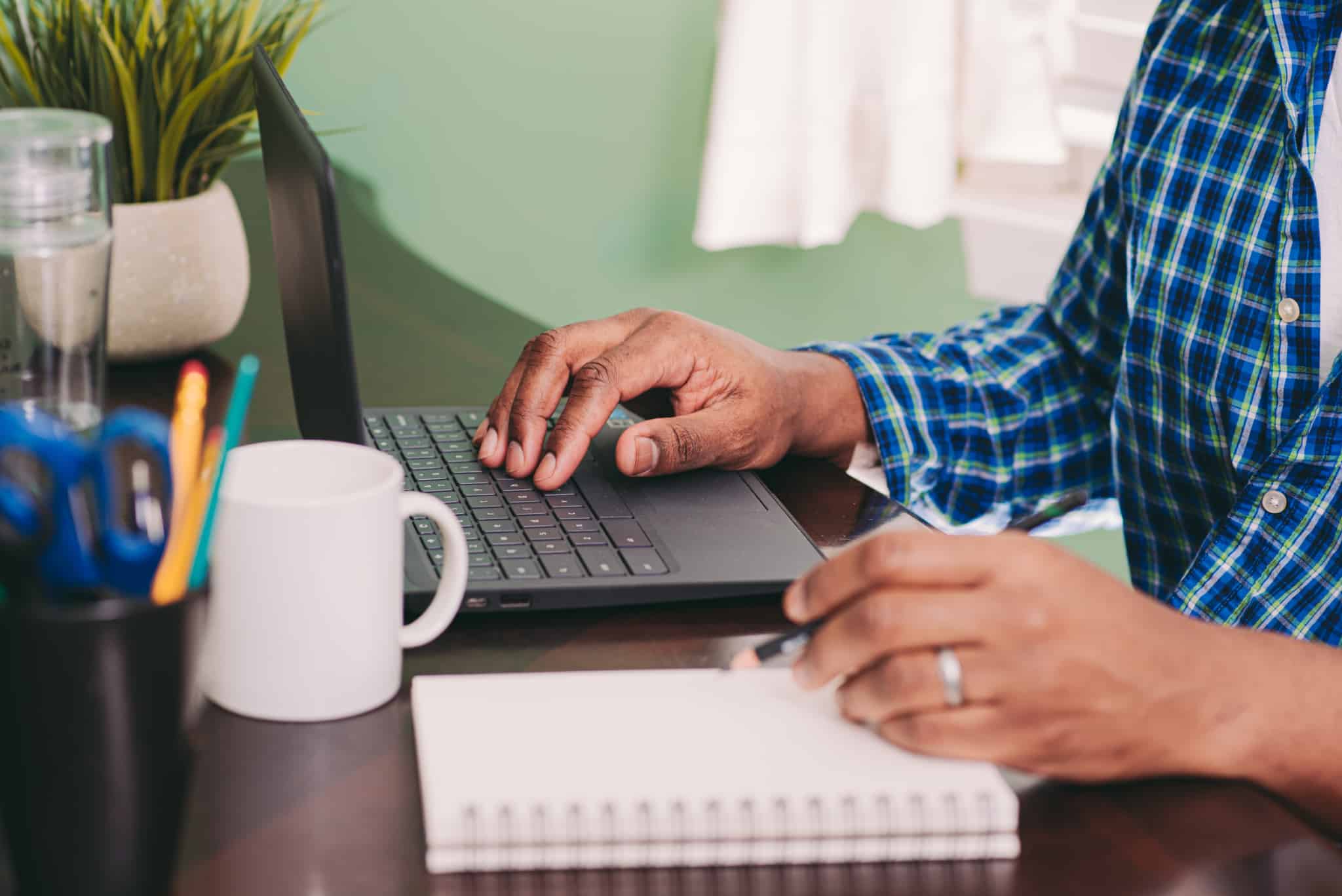 Homem com um lápis numa mão, a tirar apontamentos num caderno, enquanto consulta informação num computador
