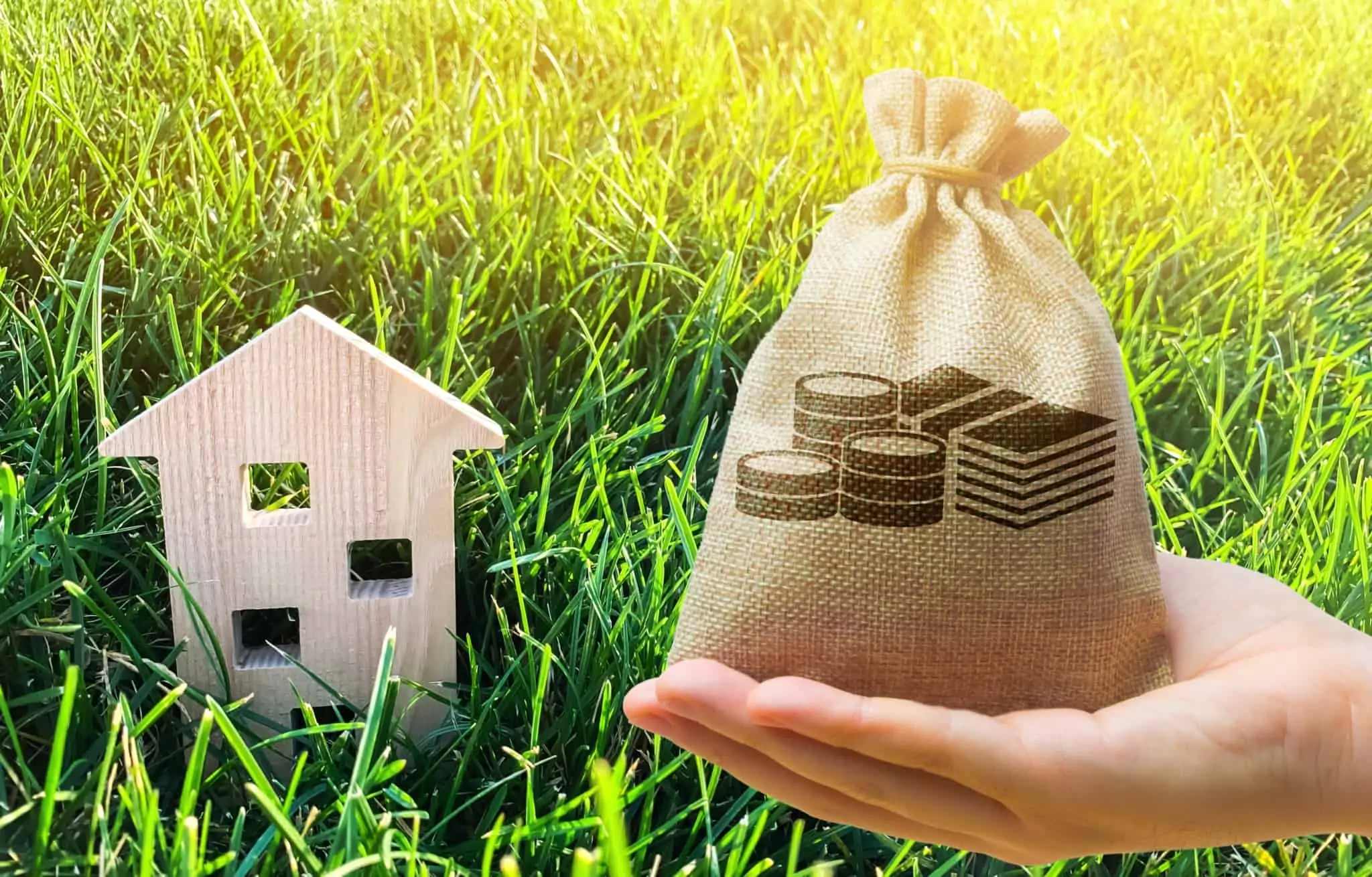 Guia de mais-valias imobiliárias Mão com um saco de serapilheira cheio de dinheiro. No plano de trás uma casa de madeira em miniatura