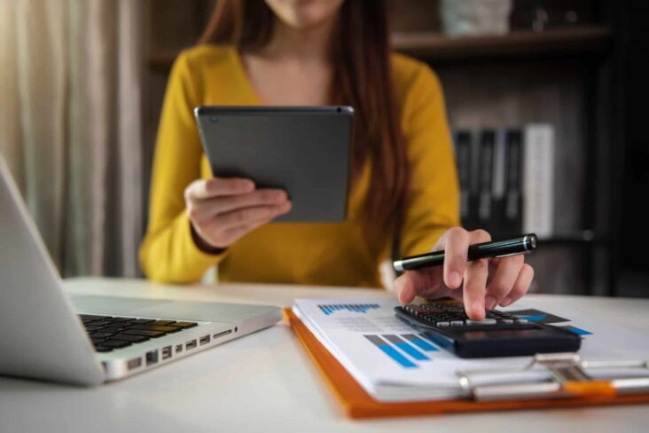 mulher faz contas com a sua calculadora enquanto confirma dados no seu telemóvel