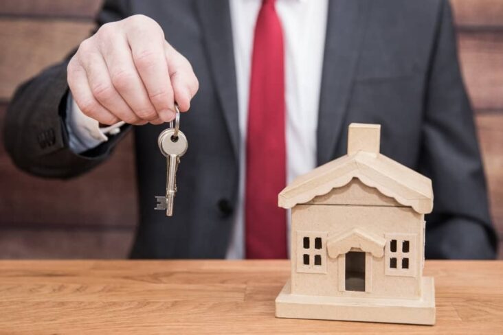 Um homem de fato e gravata sentado a entregar uma chave com uma casa pequena de madeira em cima da mesa