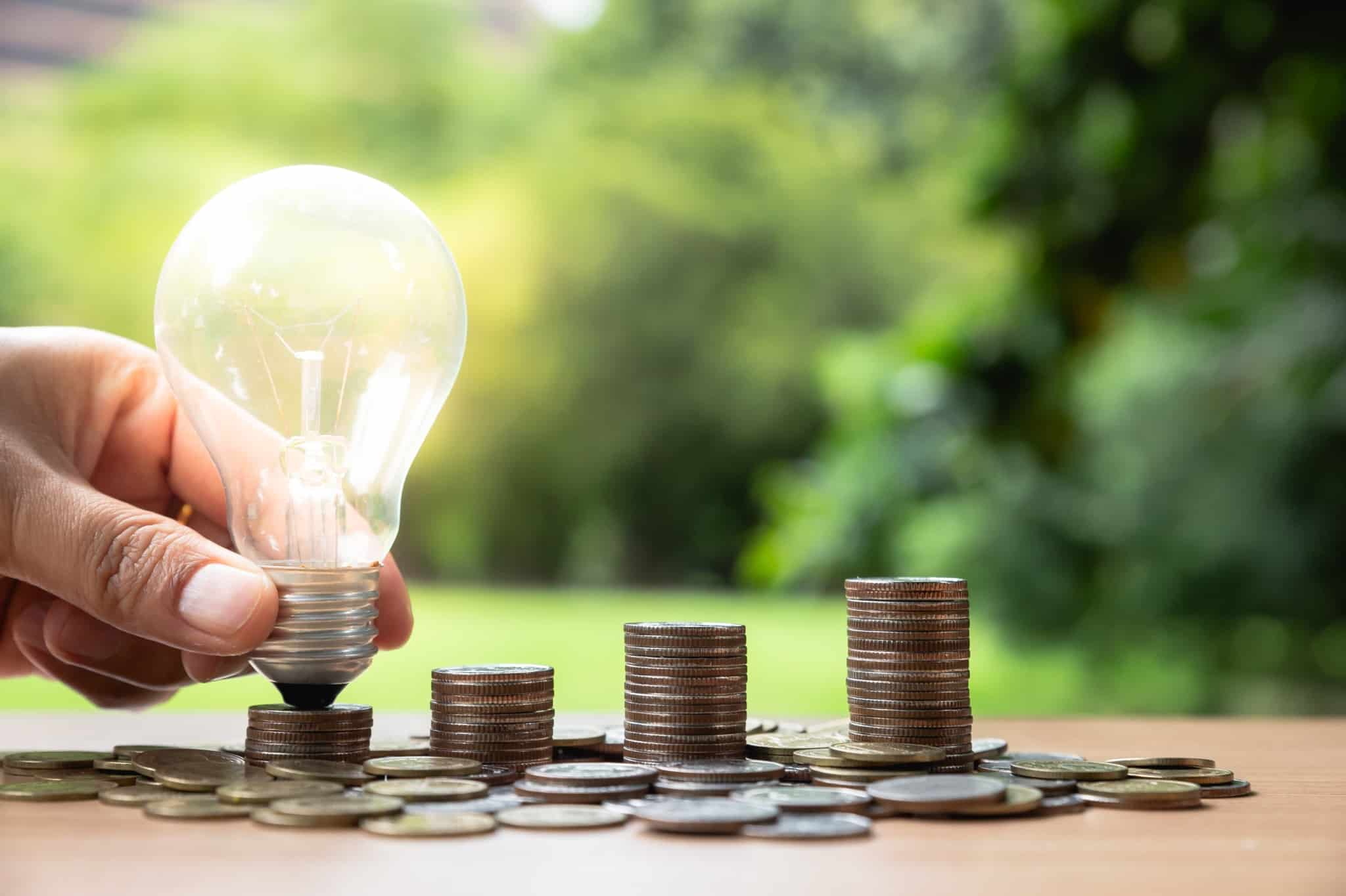 produção fotográfica a remeter para poupar na energia. Um fundo verde com natureza e quatro pilhas de moedas, numa delas uma lampada em cima, dispostas em sentido ascendente