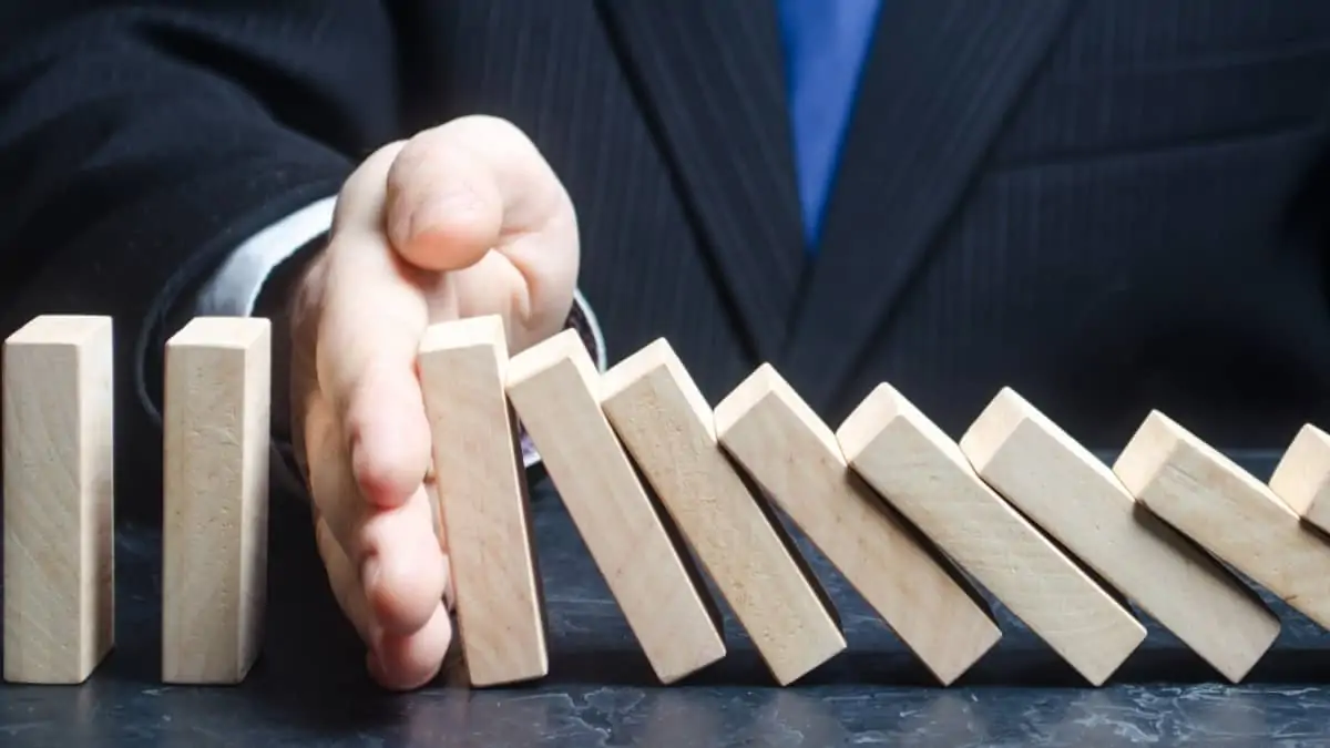 A sua empresa tem dívidas às Finanças? Saiba como pode regularizá-las Homem de fato e gravata a segurar peças de fila de dominó a cair com a mão