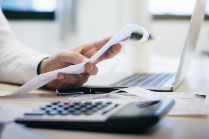 Mão de senhor a segurar um talão com uma calculadora e um portátil à frente a reequilibrar contas