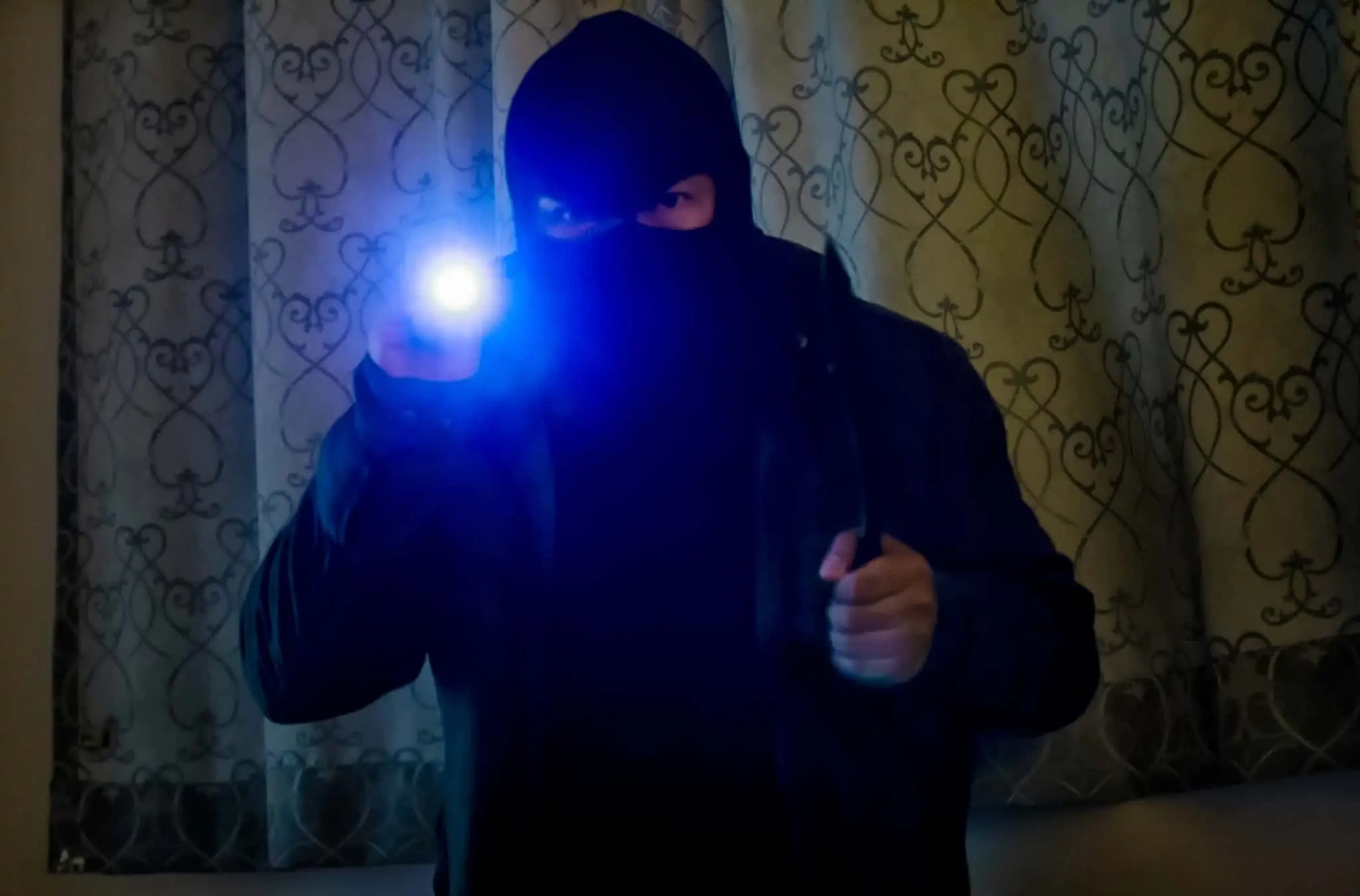 Seguro de recheio: Como funciona e quanto custa proteger a sua casa? assaltante, encapuçado, a entrar numa casa durante a noite e de lanterna na mão
