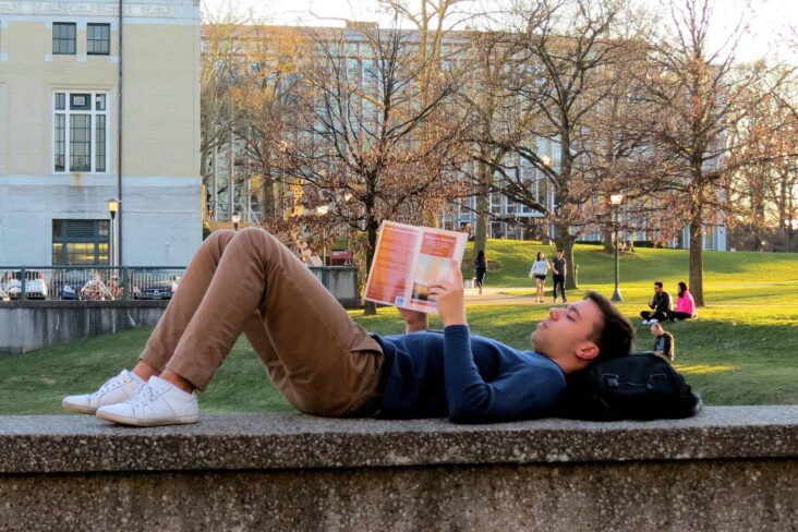 jovem estudante deitado em cima de um muro , junto à universidade, a ler um livro, com a cebça em cima da mochila