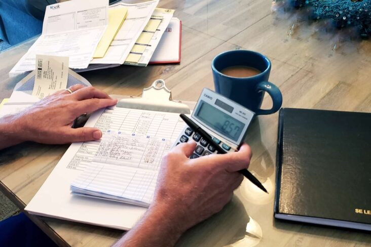 jovem homem faz calculos, recorrendo a uma calculadora, analisando um série de contas espalahdas em cima da sua mesa