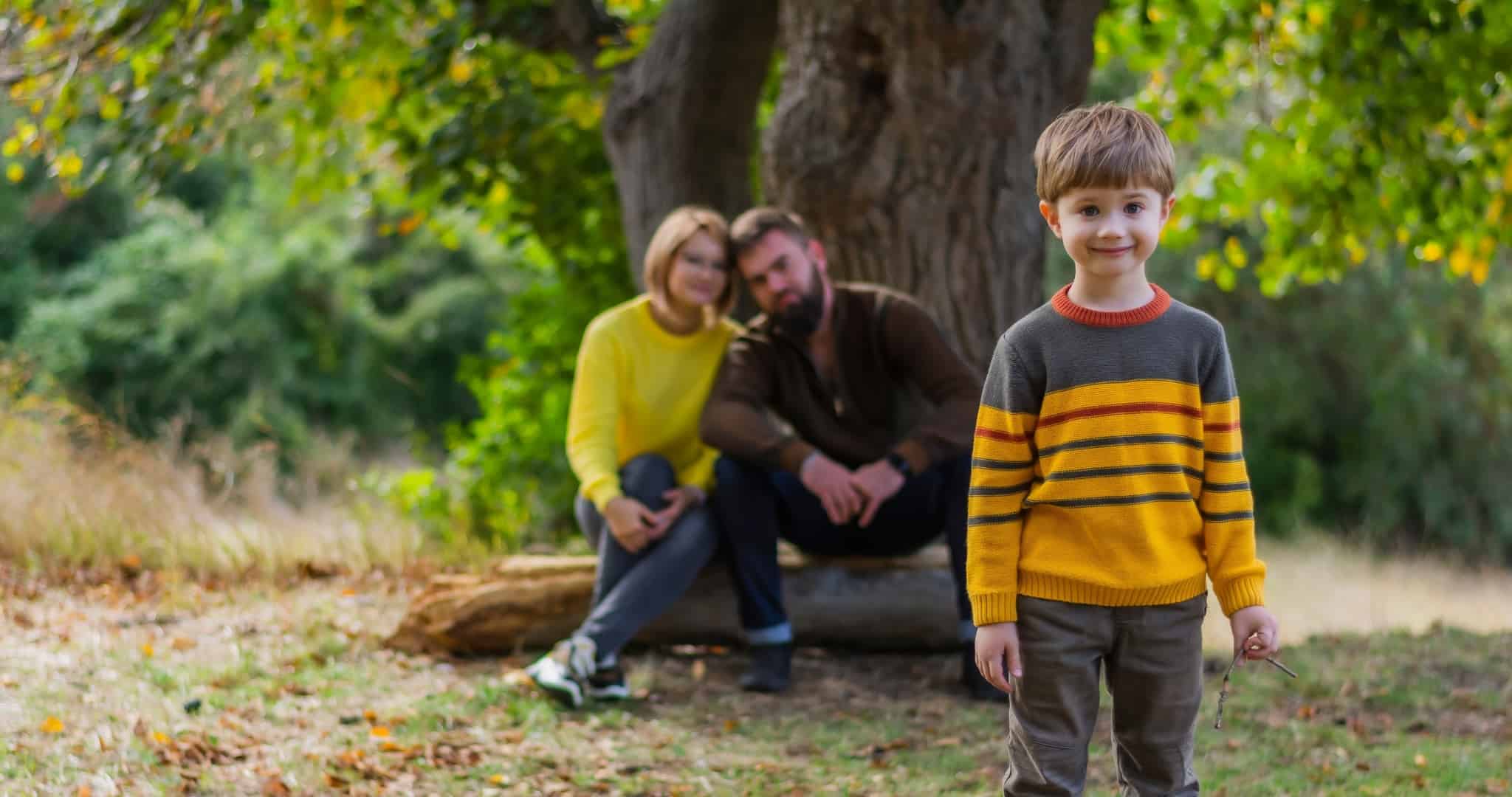 Criança adotada posa para fotografia com os pais adotivos sentados junto a uma árvore