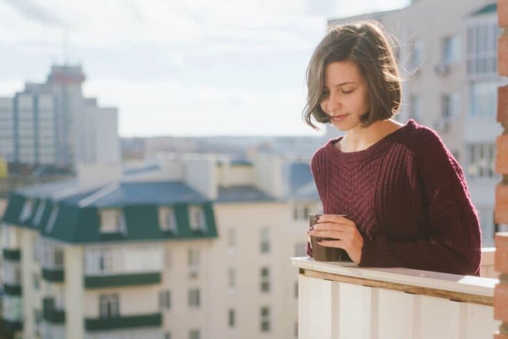 jovem mulher, de caneca de café na mão, está na varanda da casa que arrendou, a ver a vista da cidade
