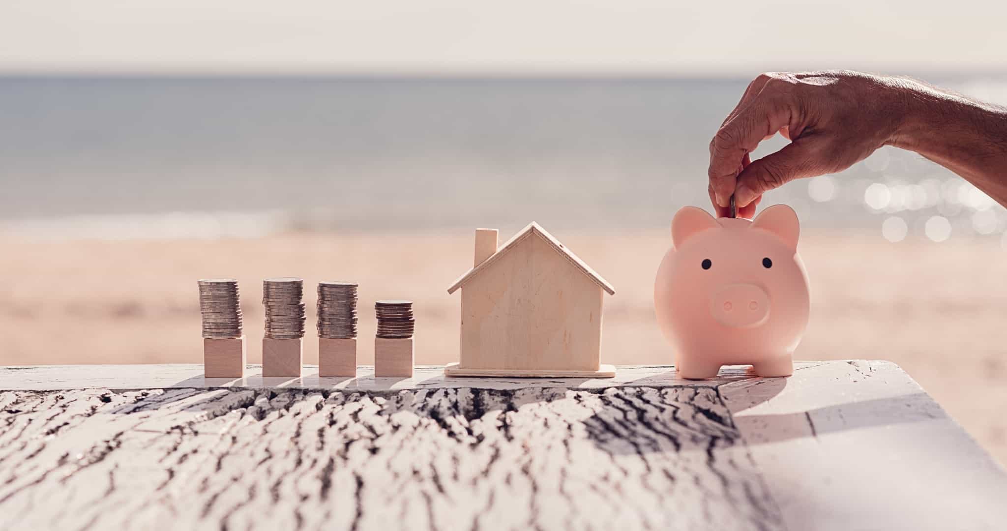 Pilhas de moedas, miniatura de casa de madeira e porco mealheiro com mão a colocar moeda lá dentro