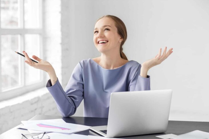 jovem mulher, sentada à secretária, festeja entusiasticamente, de braçpos levantados, ter recebido um aumento salarial