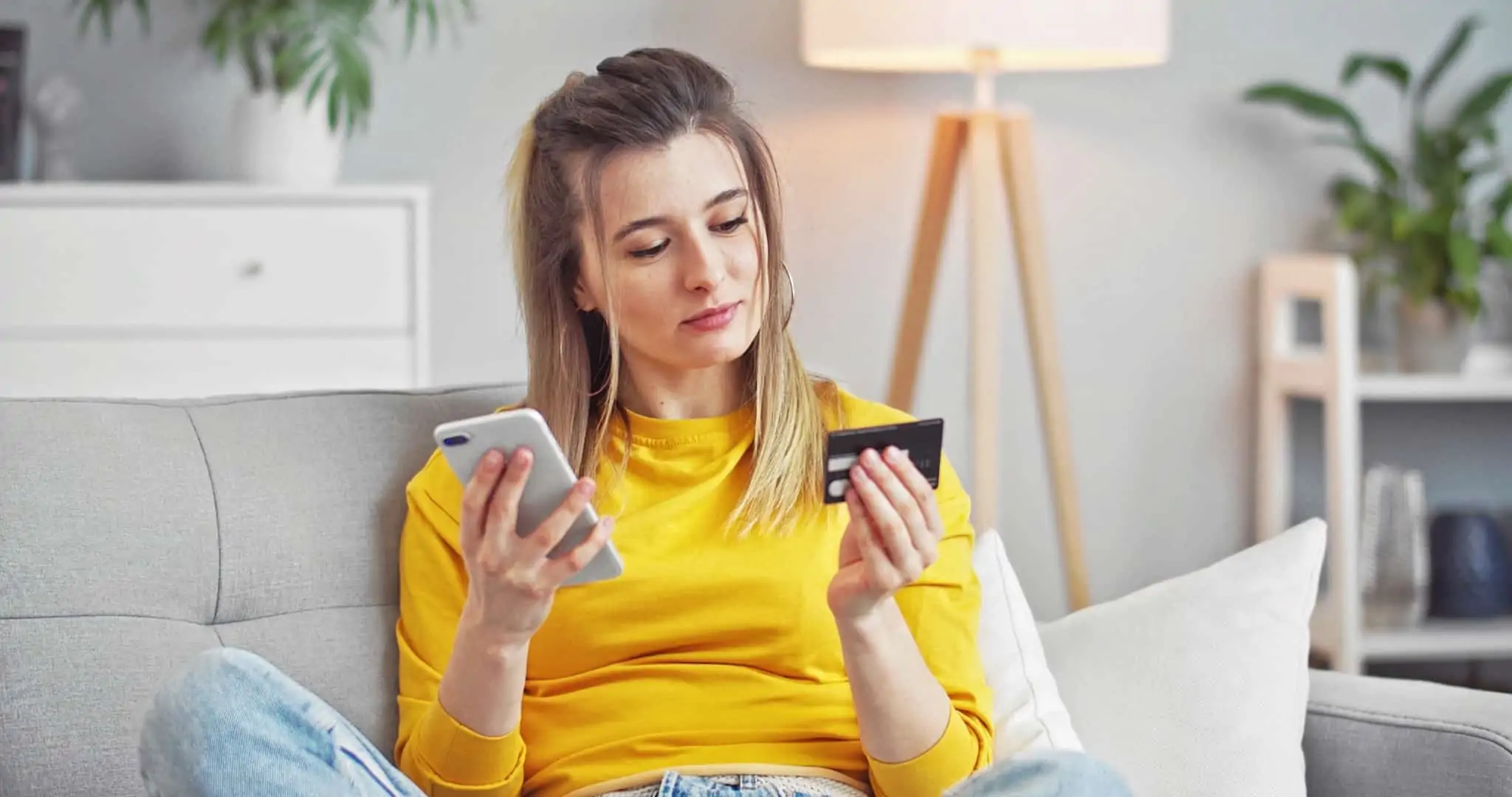 Cashback e Cash advance: Como funcionam e quais as suas diferenças? jovem mulher faz compras através do telemovel, sentada no sofá de casa, usando o cartão de crédito