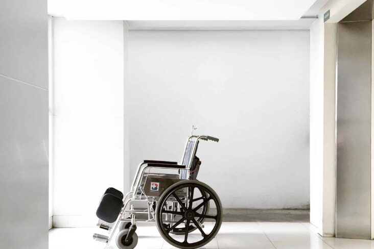cadeira de rodas, vazia, parada no meio de um corredor também vazio, branco