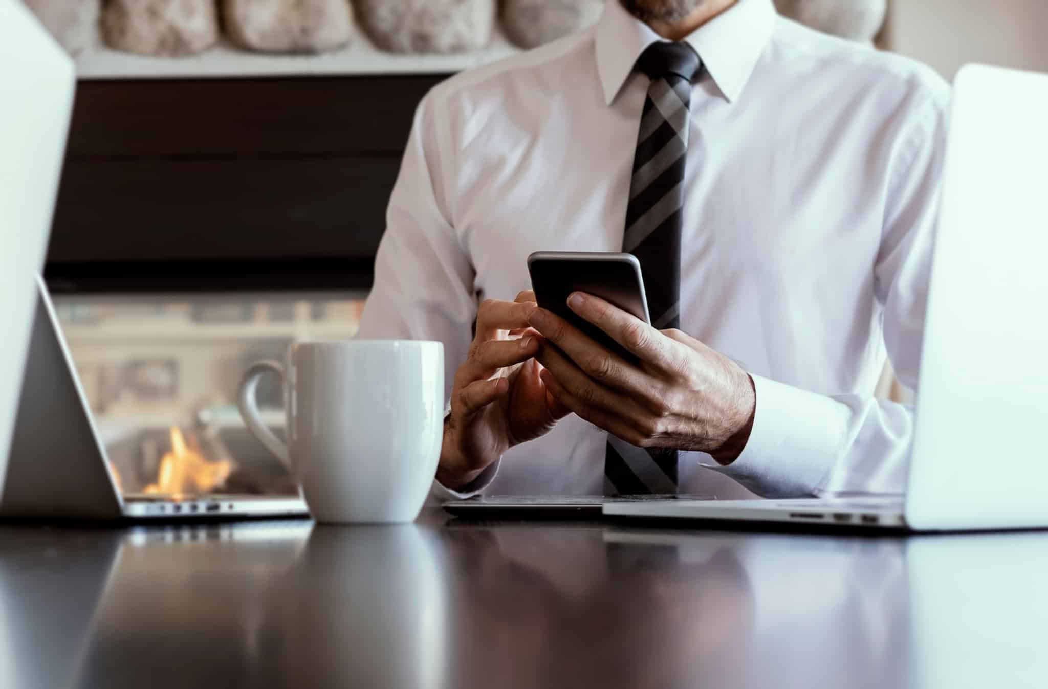 Imagem de um homem de camisa e gravata à mesa, com o telefone na mão e portáteis - escrituras à distância