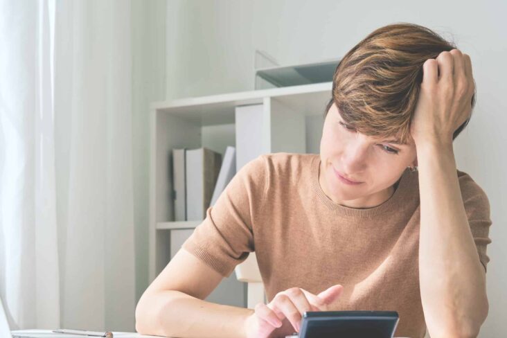 jovem mulher com a mão na cabeça e olhar preocupado faz contas, às suas dívidas, na calculadora