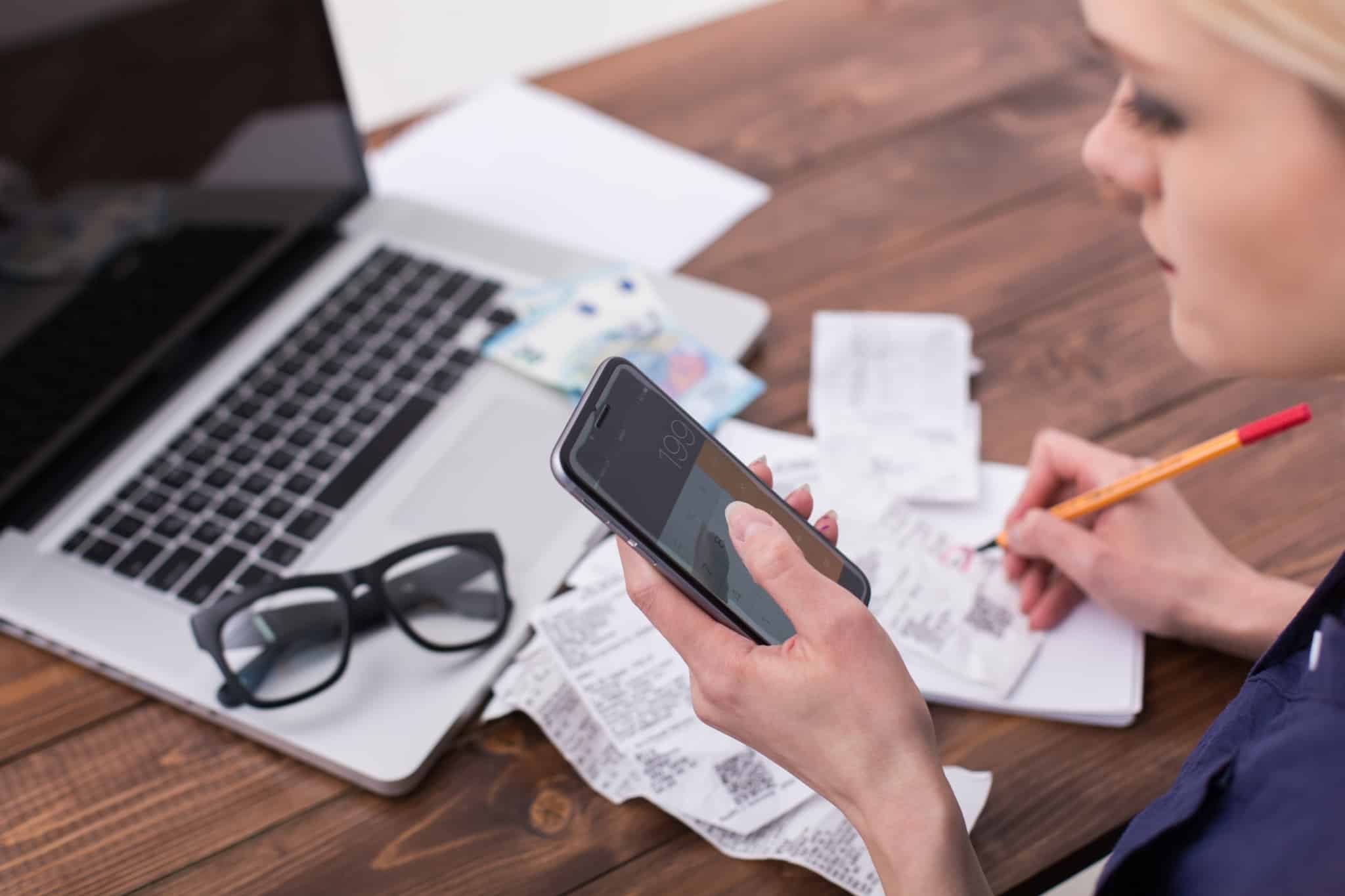 jovem mulher lista as despesas e faz contas na calculadora do telemóvel p'ara definr o seu orçamento familiar