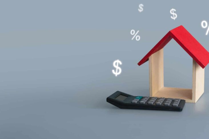 produção fotográfica mostra uma maqueta de uma casa, só com telhado vermelho e paredes laterais com calculadora ao pe e símbolos financeiros ao redor