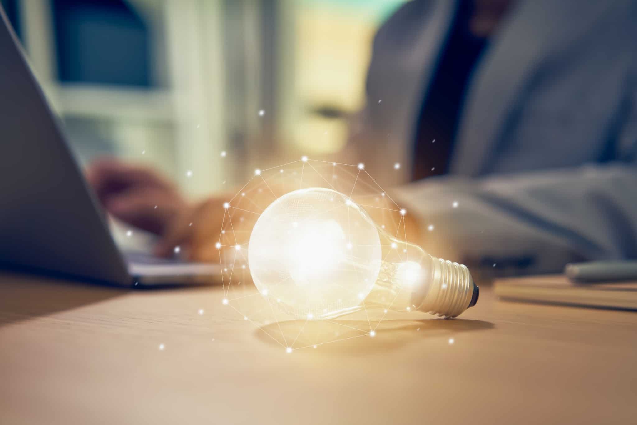 Propriedade intelectual: O que é e como funciona? com um jovem homem a trabalhar ao computador portátil, a propriedade intelectual é ilustrada por uma lâmpada acesa e brilhante em cima da mesa e junto ao homem