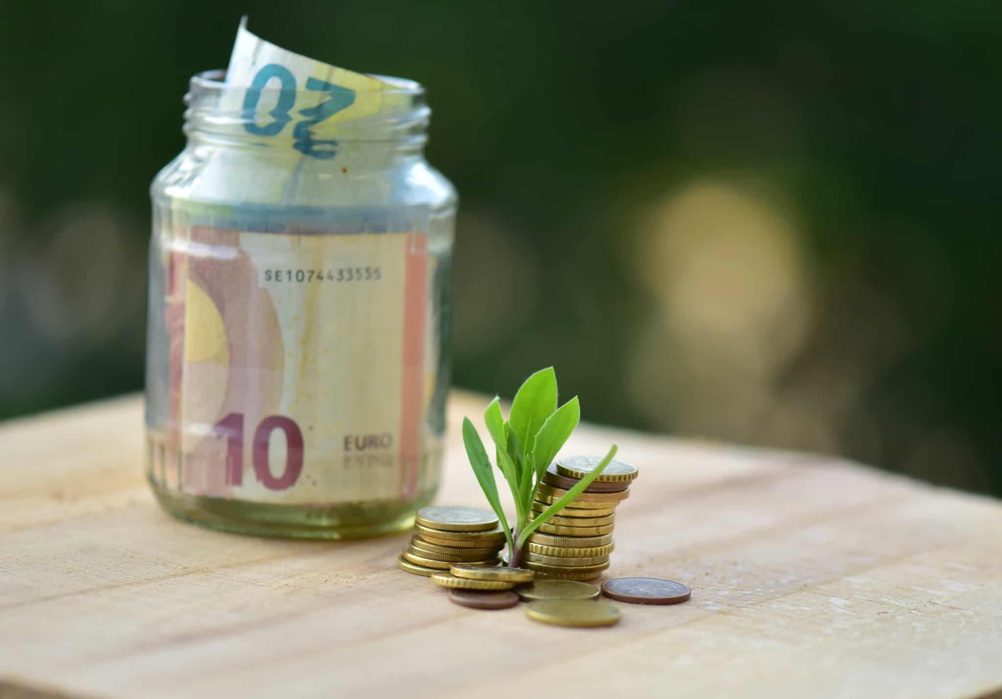 reembolso do IRS ilustrado com produção fotográfica que, num fundo desfocado, mostra uma pequeno frasco de vidro transparente com notas de 10 e 20 euros, junto a uma pequena pilha de moedas de onde brota um pezinho de uma planta