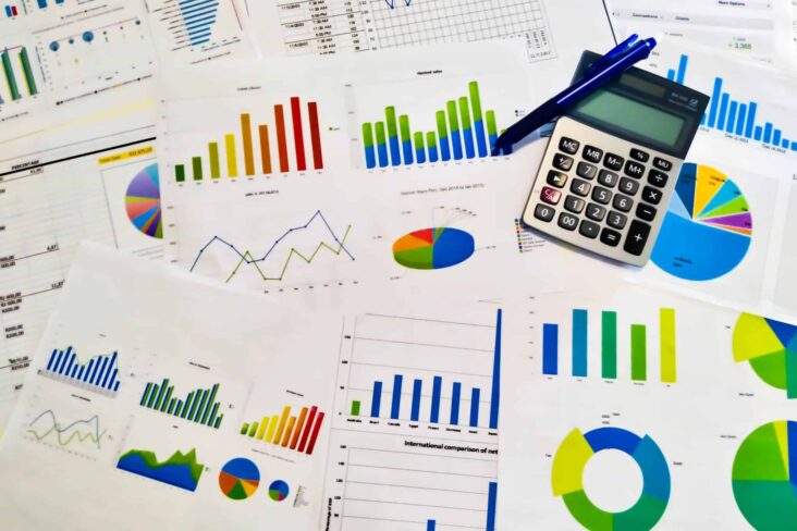 produção fotográfica que mostra um aglomerado de documentos financeiros, com gráficos de muitas cores e feitios, com uma calculadora pousada também nesta mesa, sendo que são referentes ao relatório e contas