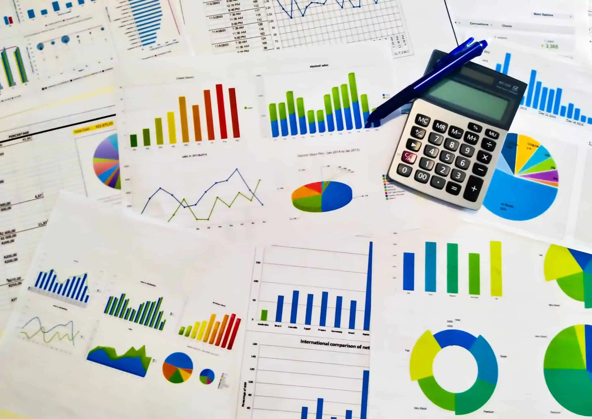 Como analisar o relatório e contas de uma empresa? produção fotográfica que mostra um aglomerado de documentos financeiros, com gráficos de muitas cores e feitios, com uma calculadora pousada também nesta mesa, sendo que são referentes ao relatório e contas