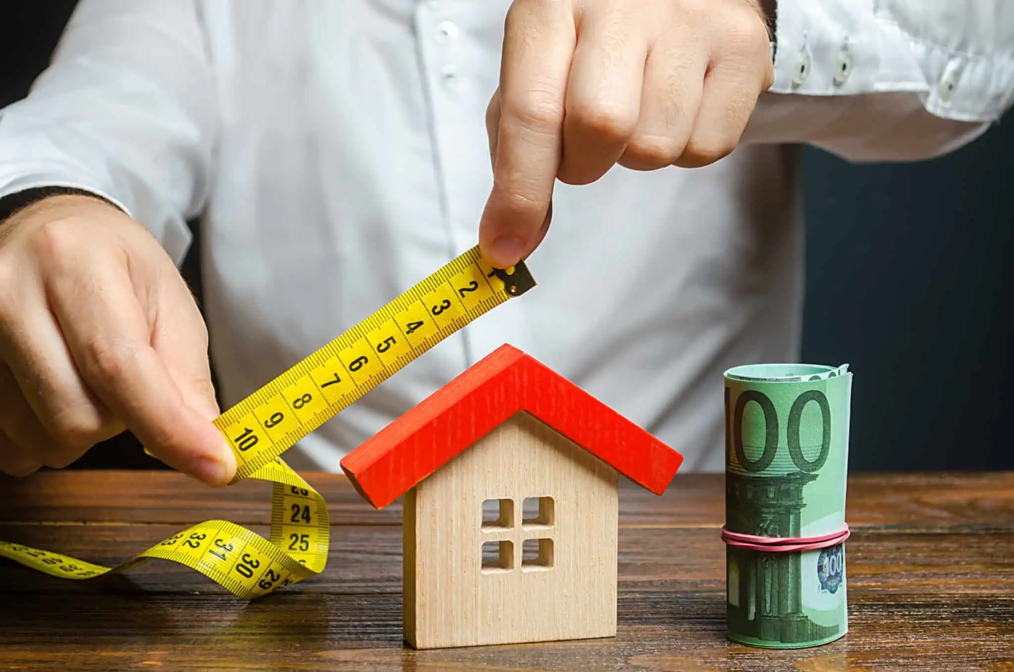 10 mitos sobre o crédito habitação um homem a medir o telhado de uma casa de madeira com um rolo de notas ao lado, servindo como referência a subida do valor dos imóveis e também aos mitos sobre o crédito habitação
