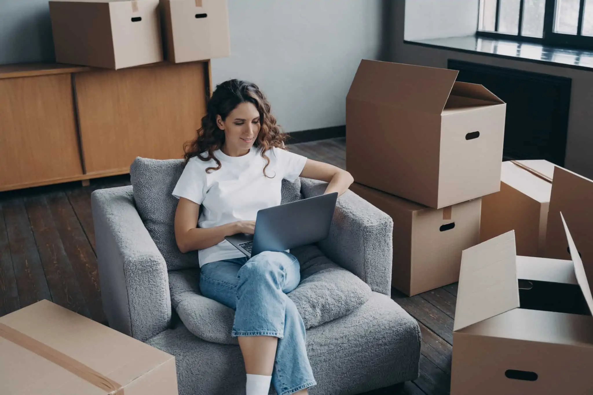 6 Sites de concursos públicos a espreitar jovem mulher, entre caixas de mudança e ao computador portátil, sentada no sofá planeia mudar de cidade por um novo emprego