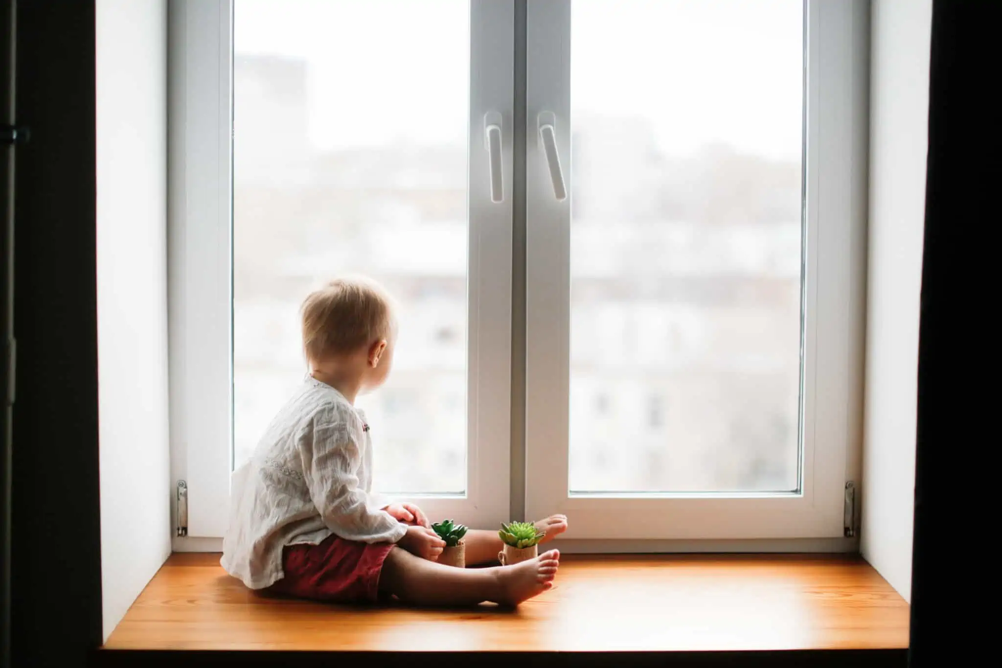 Pensão de orfandade: Como funciona? criança pequena sentada no parapeito interior de uma janela, que pode ser apoiada por uma pensão de orfandade, contempla o exterior com fraca luz do dia