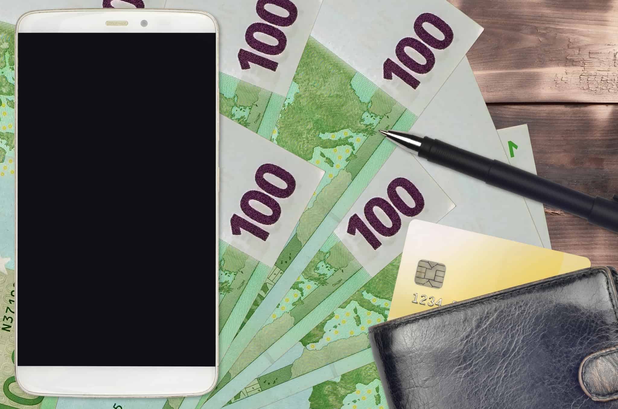 em cima de algumas notas de euros estão um smartphone branco, uma carteira da qual está a sair parte de um cartão de débito e uma centa, ilustranhdo a forma como se pode usar caixa multibanco sem cartão