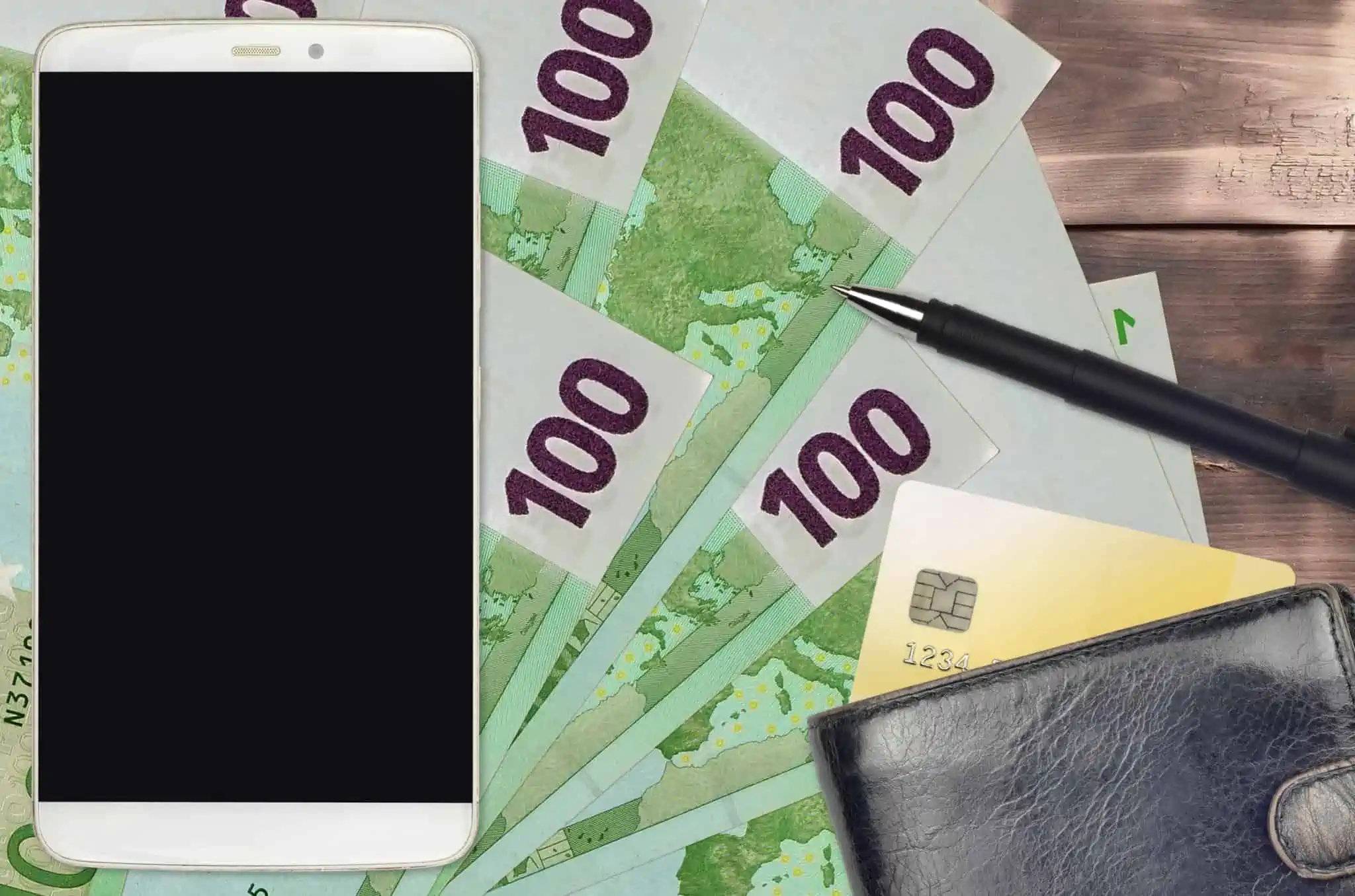 Como usar a caixa Multibanco sem cartão bancário em cima de algumas notas de euros estão um smartphone branco, uma carteira da qual está a sair parte de um cartão de débito e uma centa, ilustranhdo a forma como se pode usar caixa multibanco sem cartão