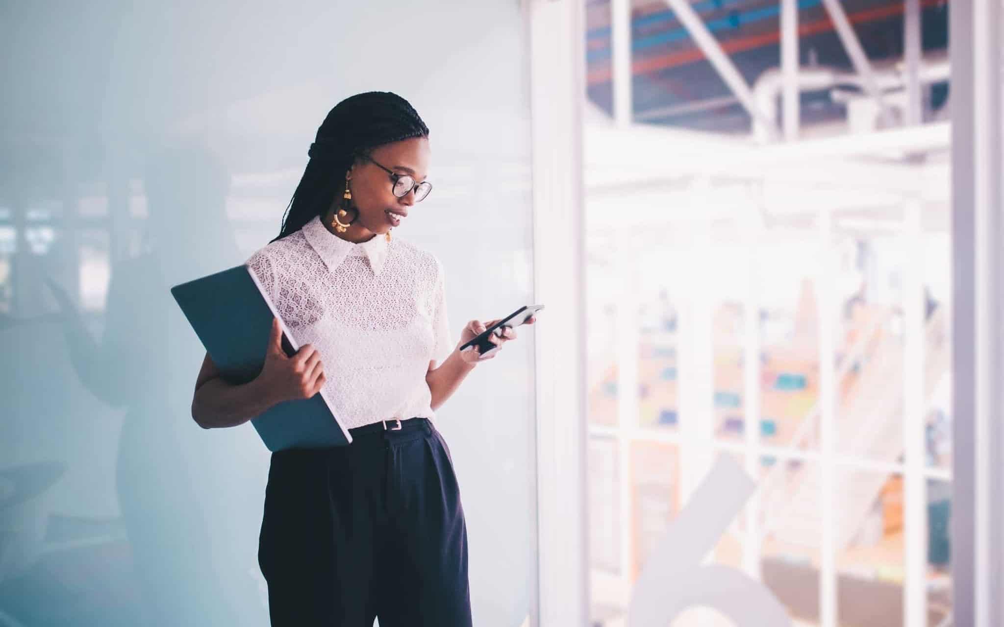 jovem mulher num escritório a segurar um telemóvel e uma pasta de documentos