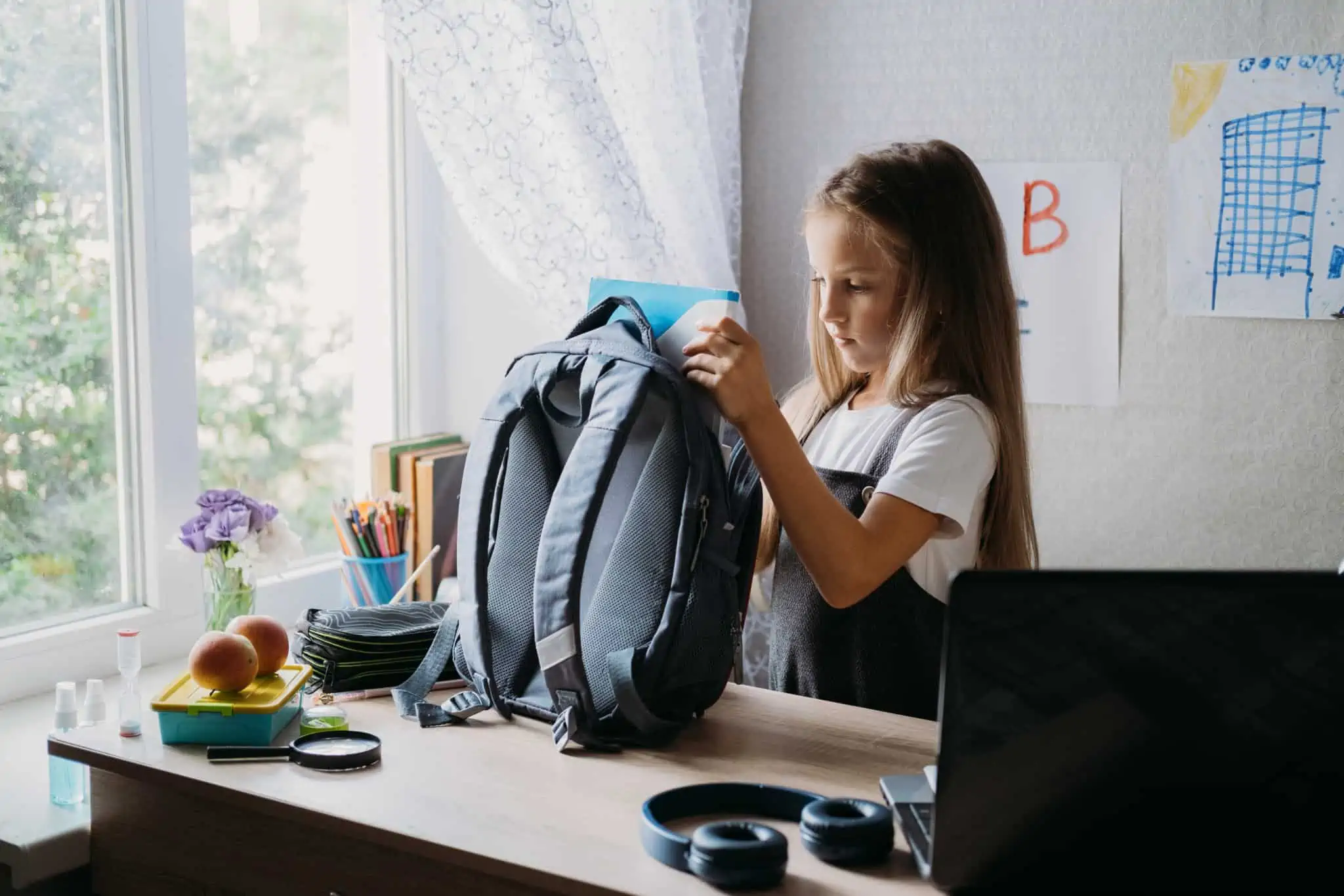 Quanto custa preparar a mochila do meu filho? Criança rapariga a colocar material escolar dentro da mochila em cima de uma mesa
