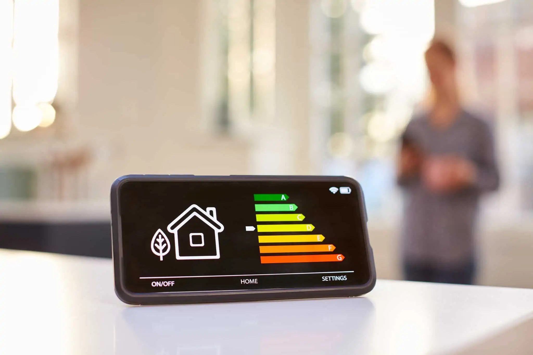 ecran de um smartphone mostra etiqueta energética de uma casa, enquanto uma pessoa conytrola essa app na sala, iliustrando o que pode ser a gestão da eficiência da energia e da água