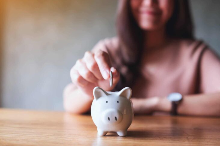 jovem mulher feliz, a sorrir, de fundo em imagem desfocada, estende a mão para colocar ima moeda no porquinho mealheiro, alimentando a poupança e o seu bem-estar