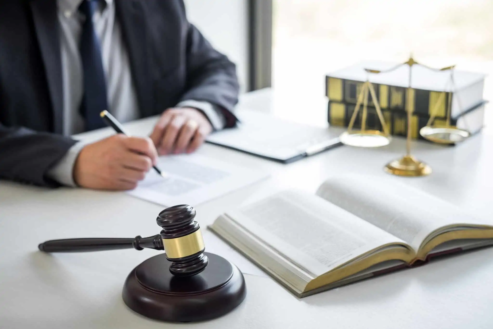 Proteção jurídica? O que é e a quem se destina homem, vestido formalmente, escreve num documento, frente a um livro aberto, exposto numa mesa com símbolos de proteção jurídica, entre eles o martelo dos juóizes e a balança da justiça