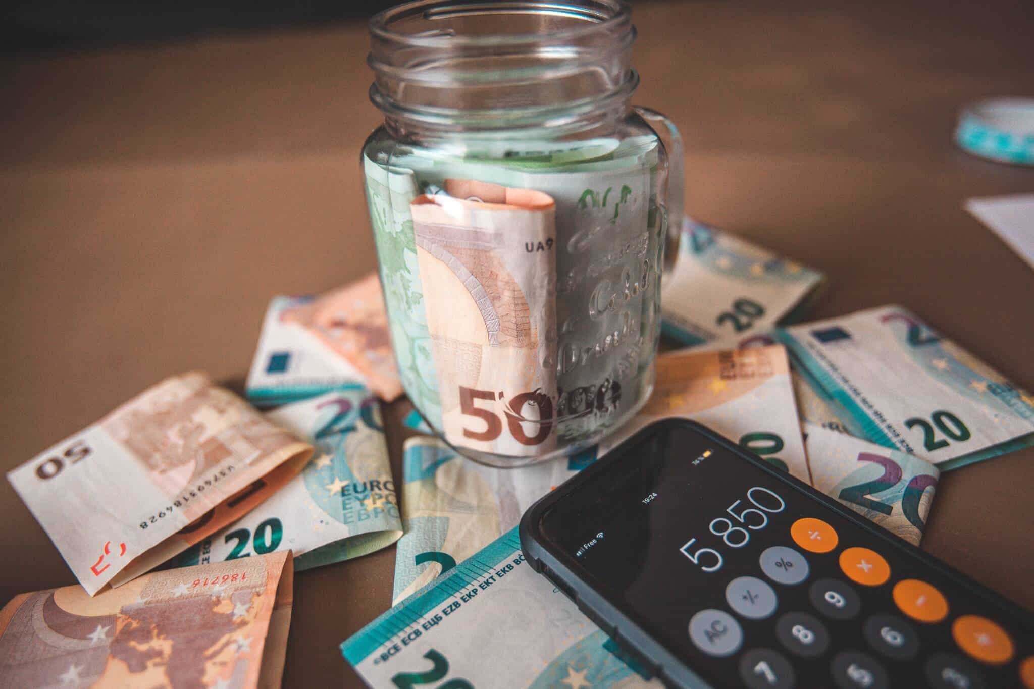Pote de vidro com notas de 50 euros e notas e telemóvel na calculadora em cima de mesa simbolizando poupança