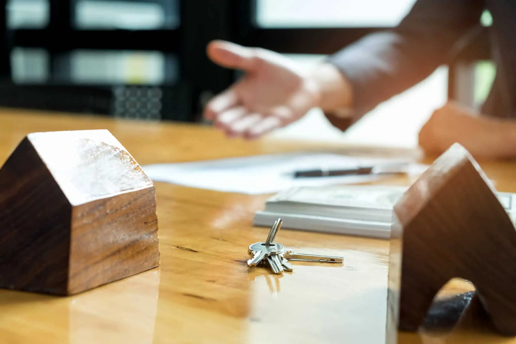 Vender imóvel herdado: Quais os passos a dar para concretizar o negócio em cima de uma mesa estão duas maquetas de casas, em madeira, junto a papeis e umas chaves de casa, num conjunto exposto por um jovem homem que explica o que fazer para vender imóvel herdado