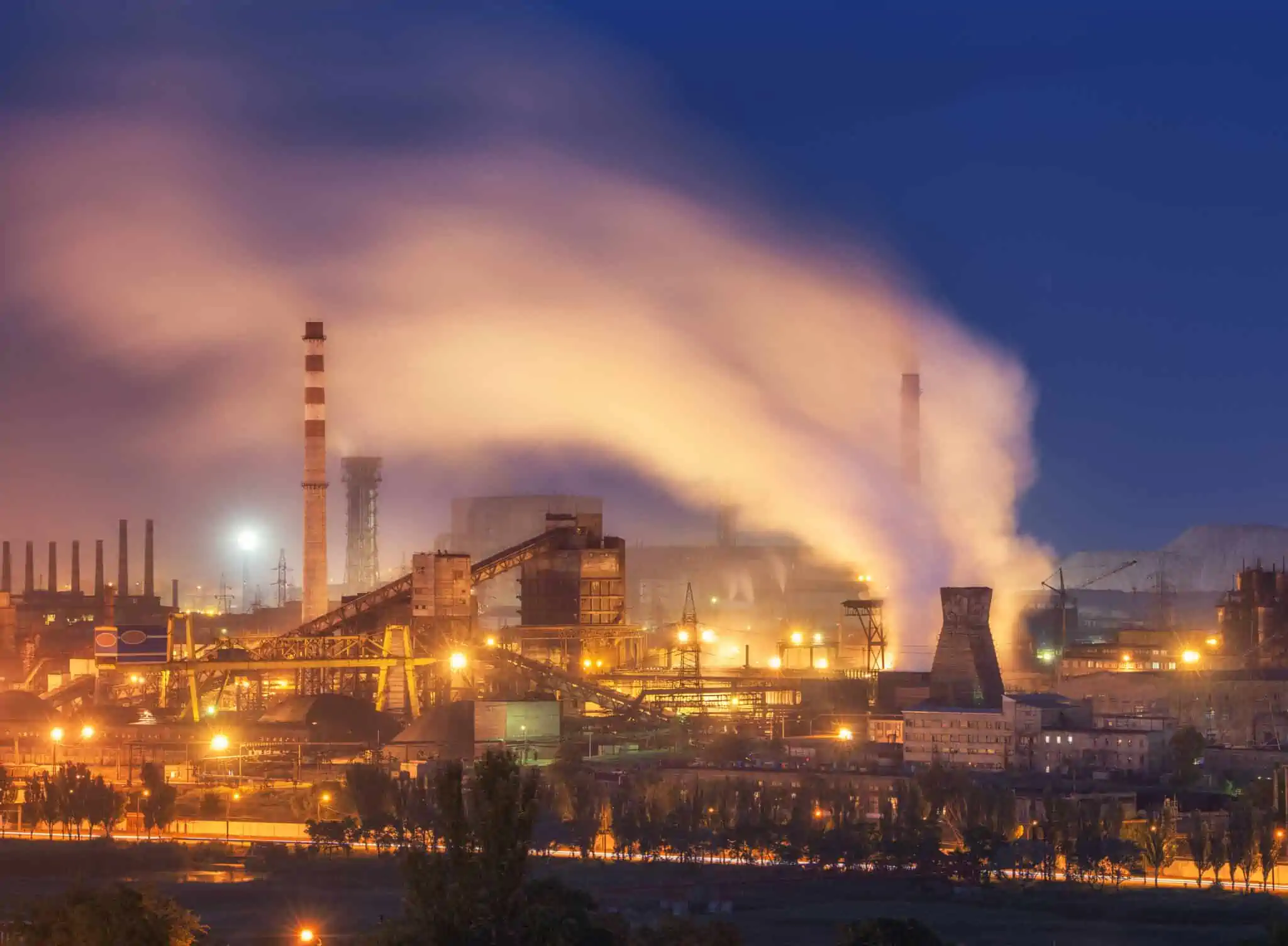 O que é e como funciona o mercado do carbono? mercado do carbono ilustrado com imagem de uma metalurgica em pleno funcionamento, durante a noite, com gfrande emissão de gases para a atmosfera