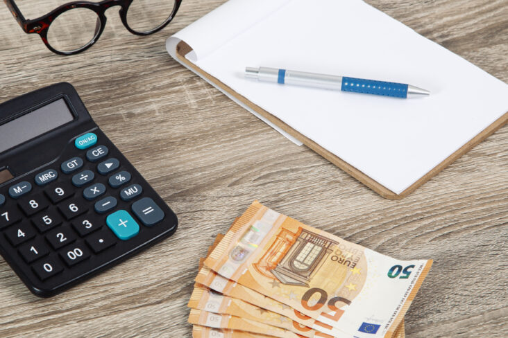 notas de 50 euros dispostas em leque em cima de uma mesa de madeira onde constam também uma calculadora, uns óculos de ver e um bloco com caneta, ilustrando assim os hábitos de poupança desejáveis