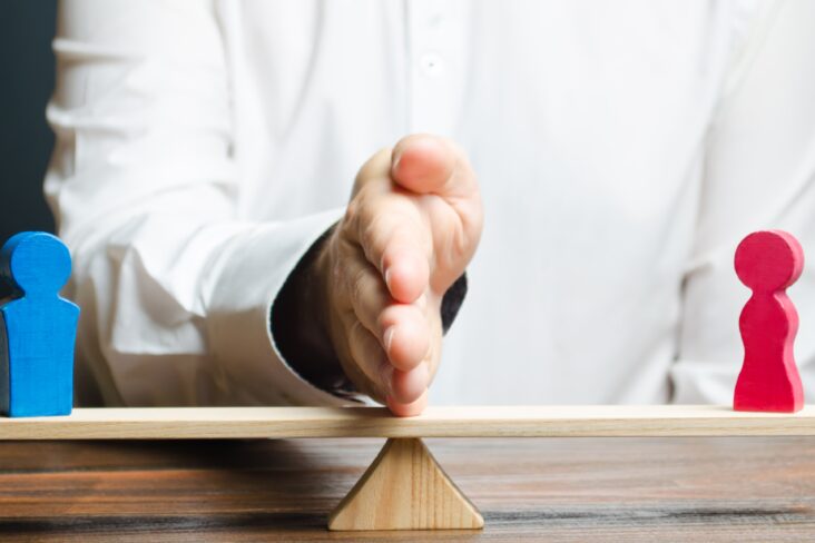 partilha de bens ilustrada com uma regua que serve de balança com um peão em cada ponta, um vermelho e outro azul, e no meio a mão de um homem, vestido de camisa branca, marca a distancia entre ambos